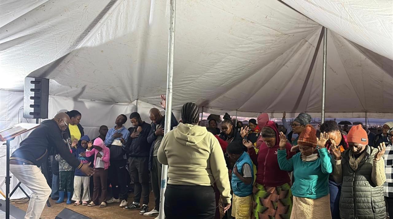 Community members praying together with raised hands under a tent